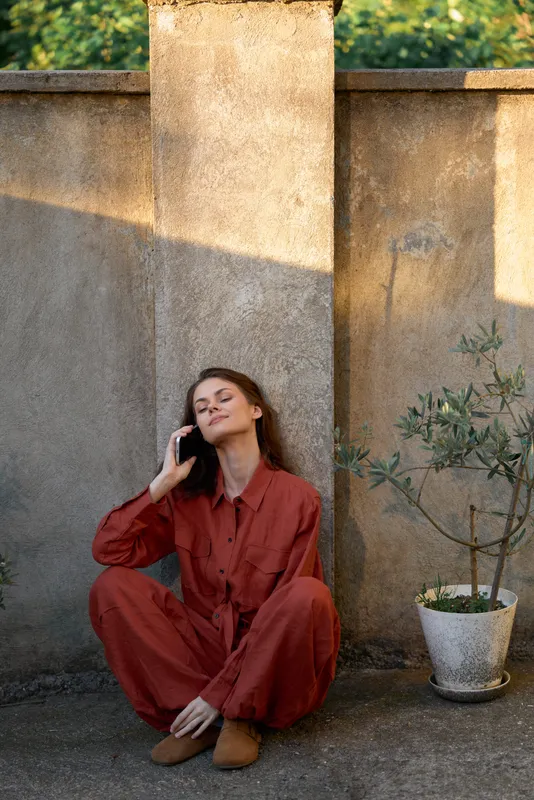 Young woman in a rust red jumpsuit sits on the ground beside a weathered wall, talking on a mobile phone with a relaxed, confident expression and natural sunlit surroundings