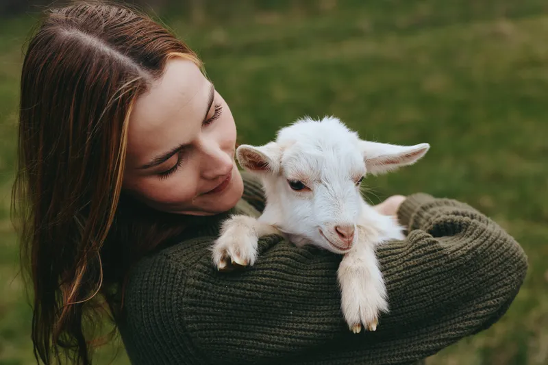 Caring woman cradles a white goat kid in her arms outdoors, soft natural light, rustic woodland mood. A wholesome lifestyle moment highlighting animal bonding, tenderness, and simple joy.