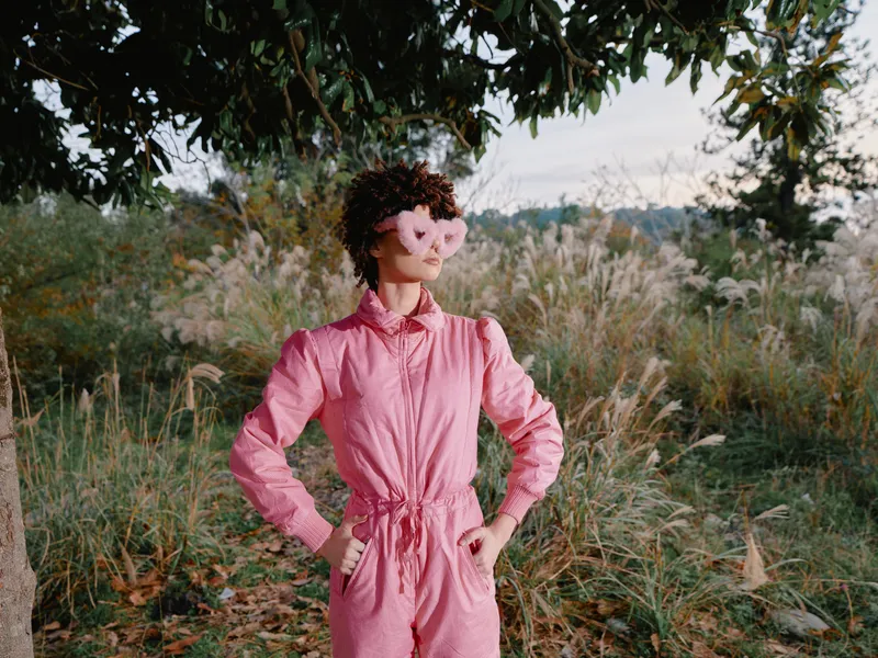 Female figure in a pink jumpsuit stands outdoors with hands on hips, wearing a heart shaped mask amid tall grasses and trees, conveying playful whimsy and bold fashion in nature.