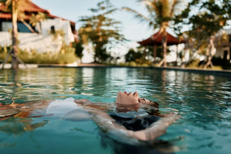 Young woman relaxing in outdoor pool on sunny day with tropical palm trees and resort buildings in background enjoying summer lifestyle and leisure time in warm weather.