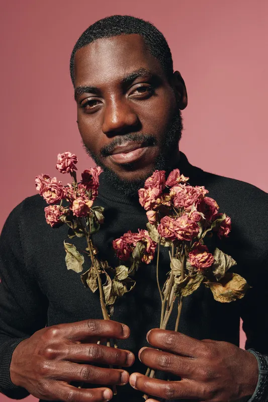 Black man with dark skin holding dried pink flowers in hands against pink background in creative visual designer style. Portrait of African ethnicity with thoughtful expression and artistic mood.