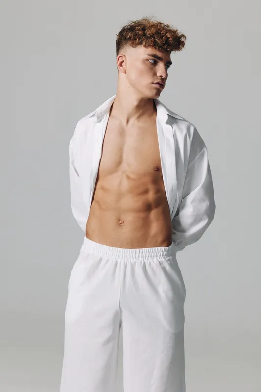 Young man posing with open white shirt and white pants in studio, showing toned abs and curly hair for fitness and fashion lifestyle concept idea.