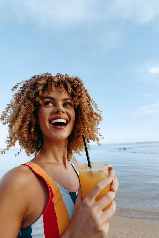 Young woman with curly hair smiles at the beach while holding a yellow cocktail, sunlit sea in the background, portraying a carefree summer vacation moment and relaxed lifestyle.