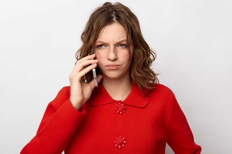 Young woman in red sweater looks worried while talking on smartphone. Isolated on white background. Concept of communication, concern, stress, and emotional reaction to news.