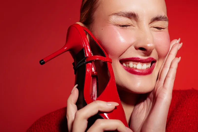 Happy woman with red lipstick holds red high heel shoe near face, eyes closed and smiling. Bright red background and clothing create vibrant, joyful mood. Concept of fashion and beauty.