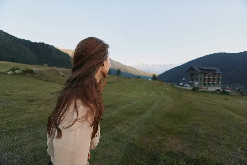 Young woman with long brown hair walks across a grassy hillside toward distant mountains and a lodge