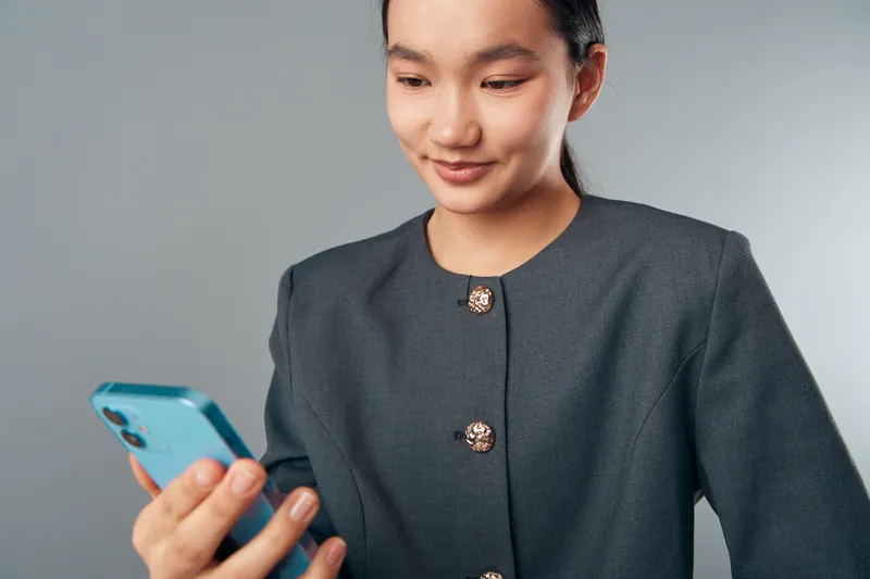 Woman in dark jacket looking at smartphone with slight smile on gray background. Female professional holding device and interacting with mobile technology in casual business style.