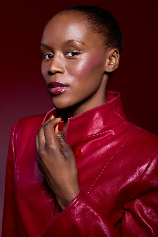 Close-up portrait of confident Black woman with dark skin wearing red leather jacket against red background. Fashion concept with bold makeup and stylish look emphasizing strength and elegance.