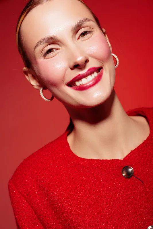 Smiling woman with red lipstick and hoop earrings wears red sweater against red background. Close-up portrait with natural skin glow and joyful expression. Concept of beauty and fashion.