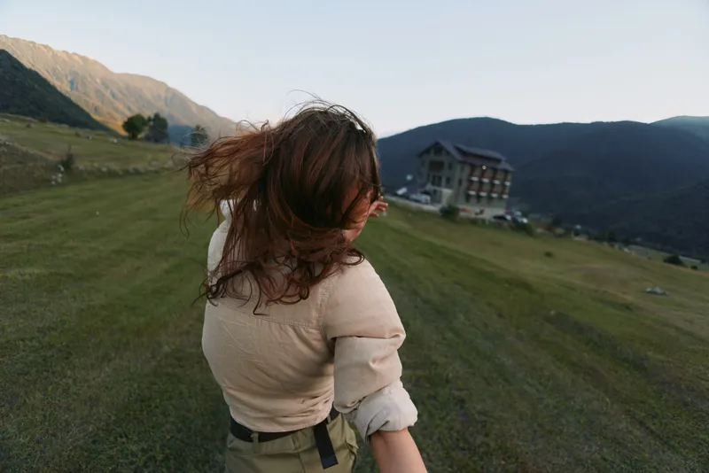 Young woman on a windy hillside with hair blown back, a distant house and mountains behind her, conveying freedom and outdoor lifestyle.