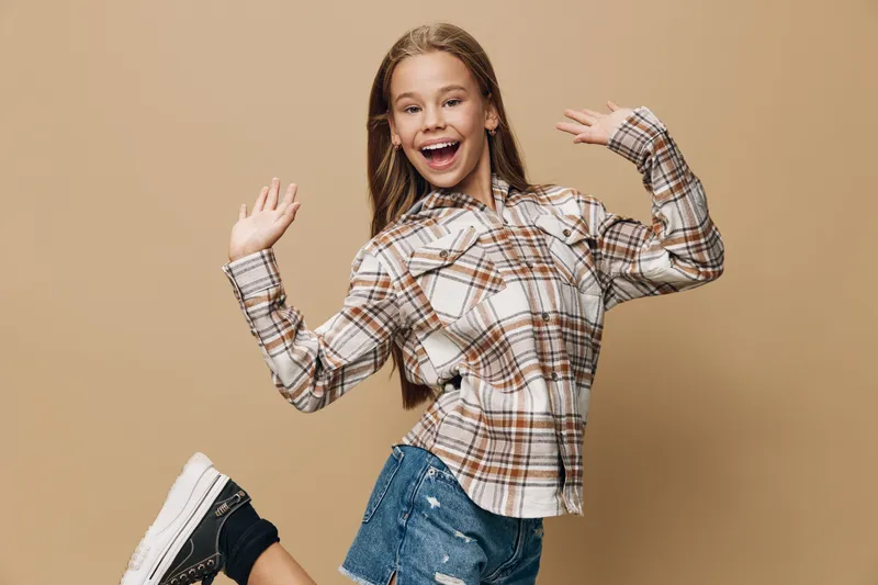 Girl in plaid shirt and denim shorts jumping with joy on beige background, smiling and looking happy with energetic pose and casual style.
