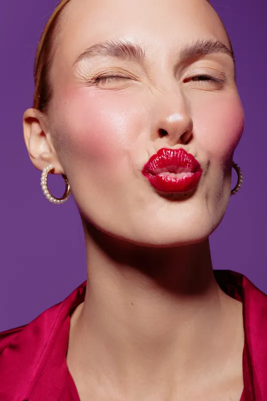 Young woman with glossy red lipstick puckers lips and winks. She wears pearl earrings and a red satin blouse against a purple background. Concept of beauty, makeup, and playful expression.