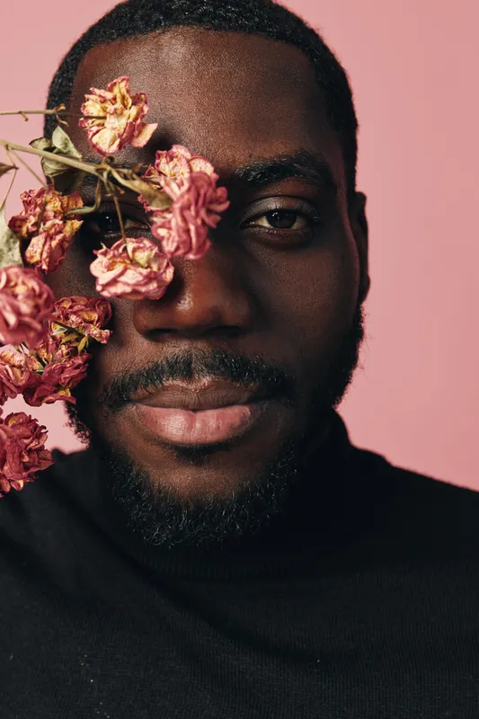 Creative portrait of African man with dark skin and beard holding dried pink flowers near face against pink background. Conceptual visual style with closeup and artistic expression in studio.