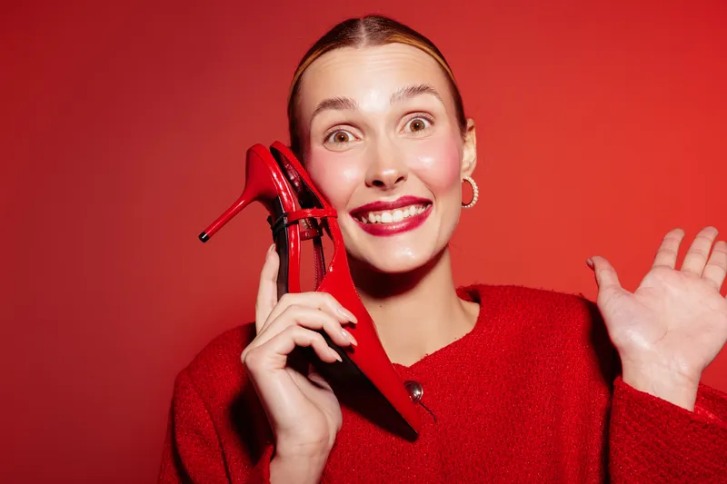 Smiling woman in red sweater holds red high heel shoe near face, posing on red background. Concept of fashion, style, happiness, and vibrant color contrast.