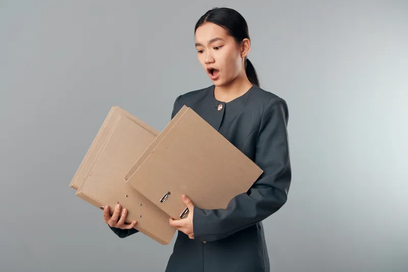 Woman holding multiple cardboard folders with surprised expression, office worker managing documents, files and paperwork with gray background and professional outfit indoors.