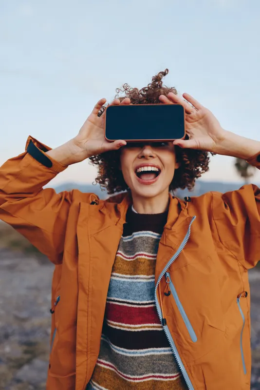 Woman, smartphone, lifestyle, laughing, outdoors, curly hair, orange jacket, holding phone above head, sharing a joyful moment, bright blue sky, candid portrait of communication