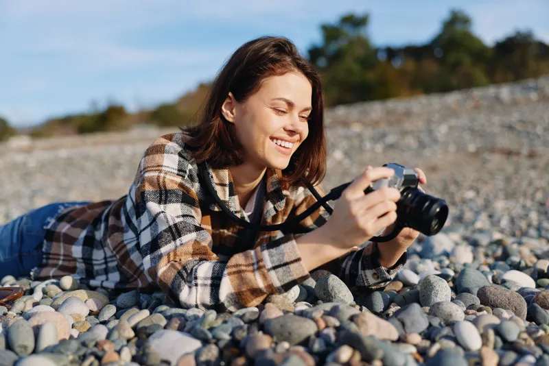Female photographer lying on a pebble beach, smiling while shooting with a DSLR, wearing a plaid shirt, enjoying nature and outdoor photography, capturing candid moments and sea scenery with creative