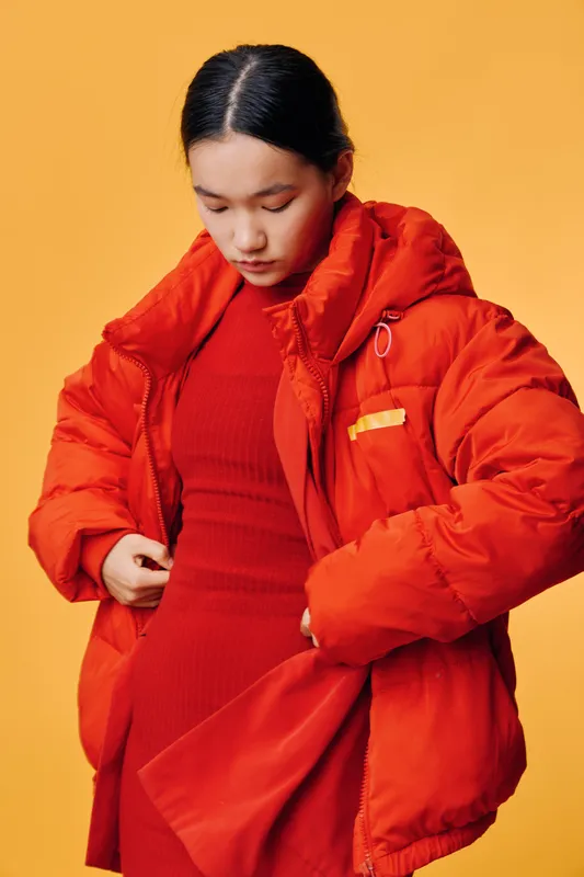 Young woman wearing red dress and oversized orange puffer jacket standing against yellow background, adjusting her coat with serious expression and casual hairstyle.