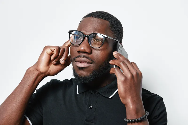 African American man with dark skin and glasses talking on smartphone, wearing black polo shirt and bracelet, isolated on white background with thoughtful expression.