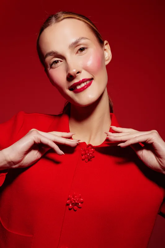 Smiling woman in red sweater poses with red beaded necklace on red background. Close-up portrait with bright makeup and natural skin glow. Concept of fashion, beauty, and elegance.