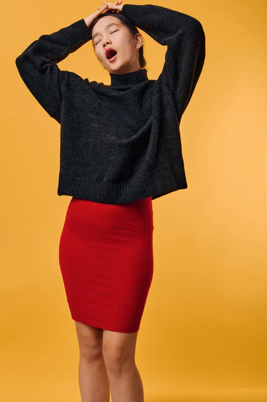 Young woman yawning and stretching with eyes closed wearing black sweater and red skirt standing against yellow background in casual fashion outfit portrait.