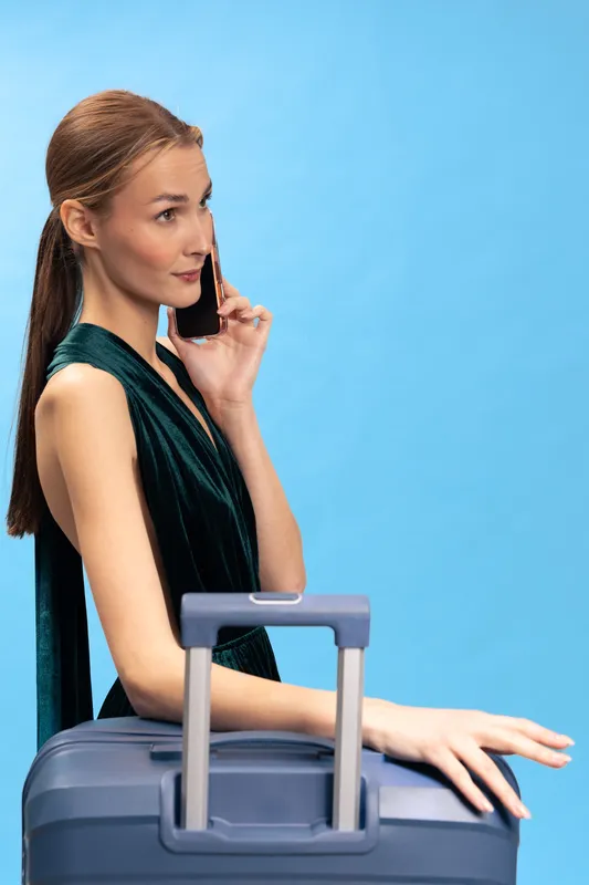 Young woman in black dress talks on smartphone while standing with suitcase on blue background. Concept of travel, business trip, and communication.