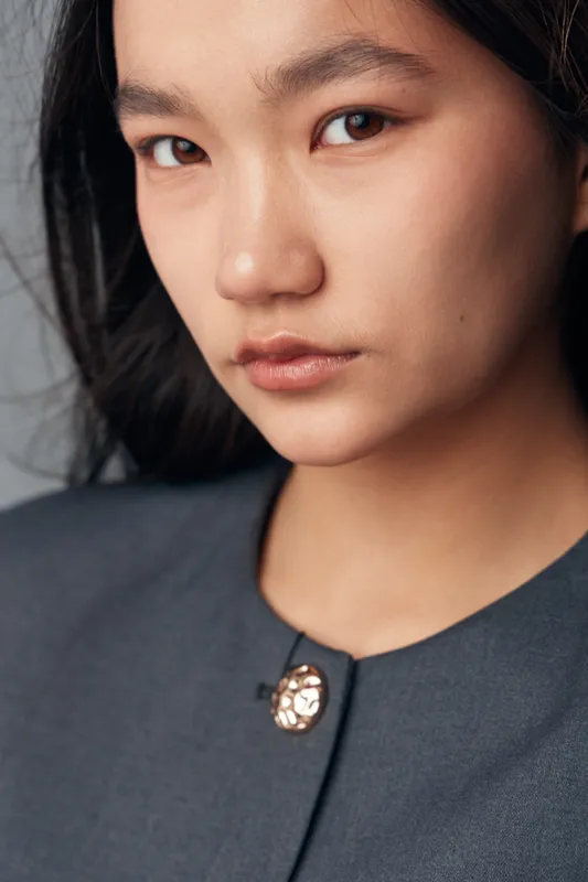 Closeup portrait of confident young woman with natural skin and intense gaze wearing gray outfit with decorative button. Serious expression and smooth dark hair detail on neutral background.