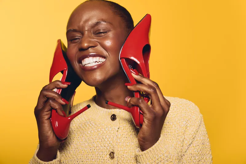 Happy African woman with dark skin smiles holding red high heels near face. Yellow background and beige sweater create cheerful mood. Concept of fashion, joy, and beauty with vibrant colors.