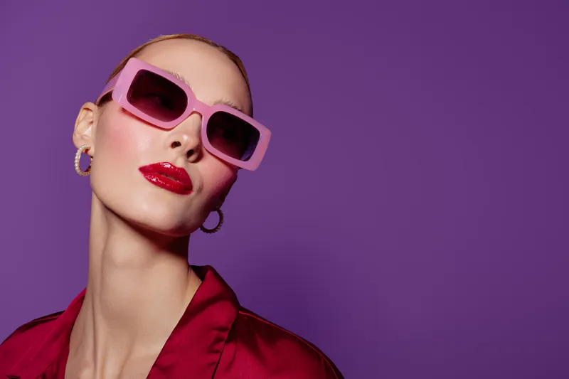 Stylish woman with pink sunglasses and red lipstick poses against purple background. Fashion portrait with bold makeup and trendy accessories. Concept of modern style and confidence.