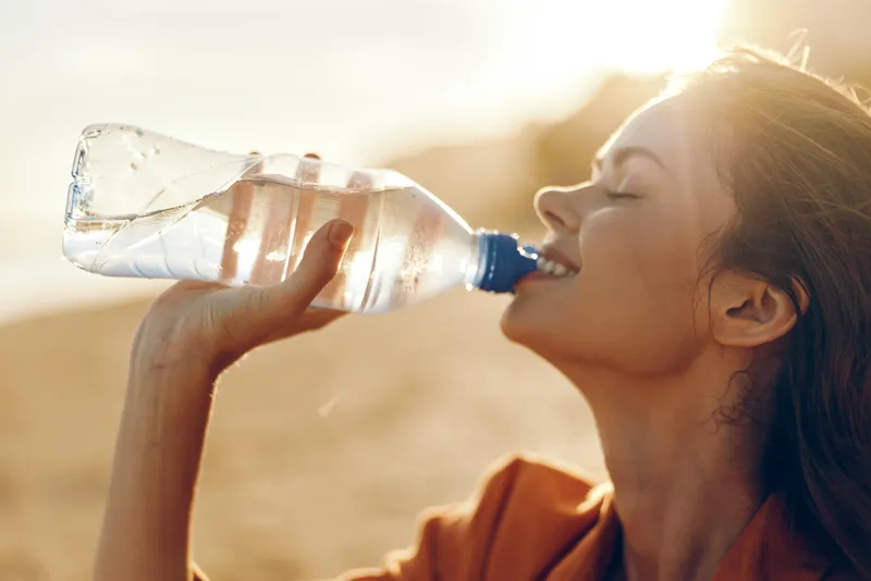 Close up profile of a healthy woman drinking water from a bottle outdoors in warm sunlight, summer hydration and refreshment concept, natural wellness drink after exercise