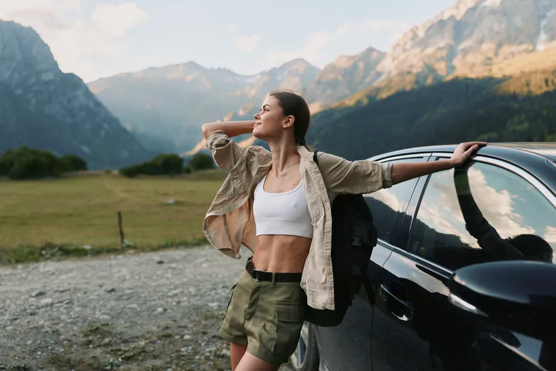 Young woman outdoors leans against a car in a mountain valley, wearing a white crop top and olive shorts, enjoying fresh air and sunshine with a carefree pose