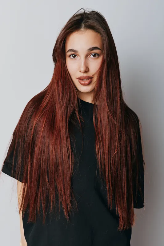 Young woman with long straight red hair wearing black t shirt standing against white background. Portrait of female model with natural makeup and casual style looking at camera.