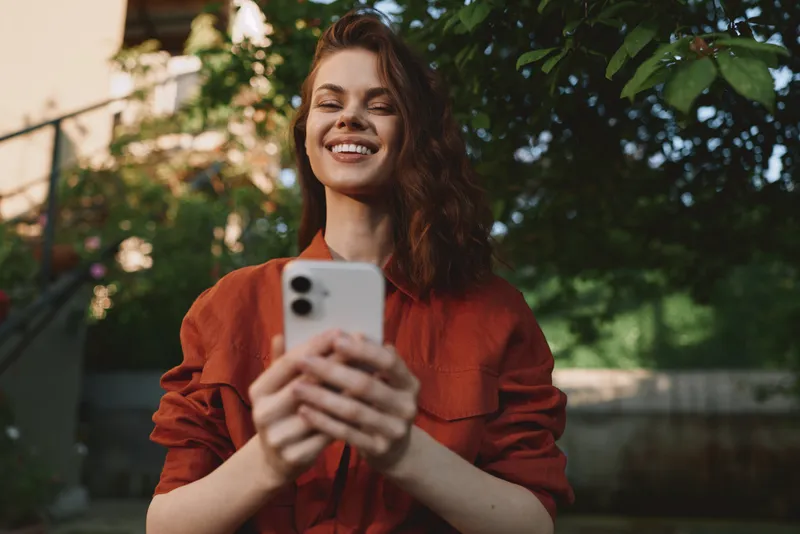 A smiling young woman in a red blouse holds a smartphone outdoors, capturing a candid moment of everyday confidence and online connection in a sunlit garden
