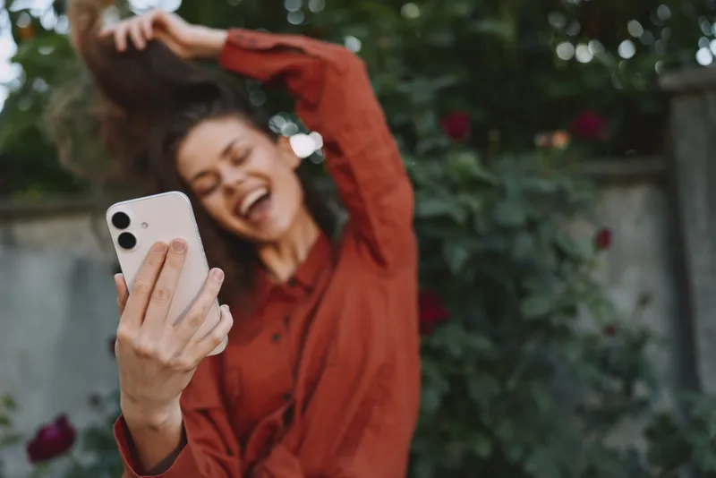 Young woman in a red blouse takes a joyful selfie outdoors, smiling broadly as she lifts her arm to capture a candid moment among lush greenery