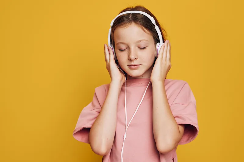Young girl in pink shirt listens to music with headphones with closed eyes on yellow background.