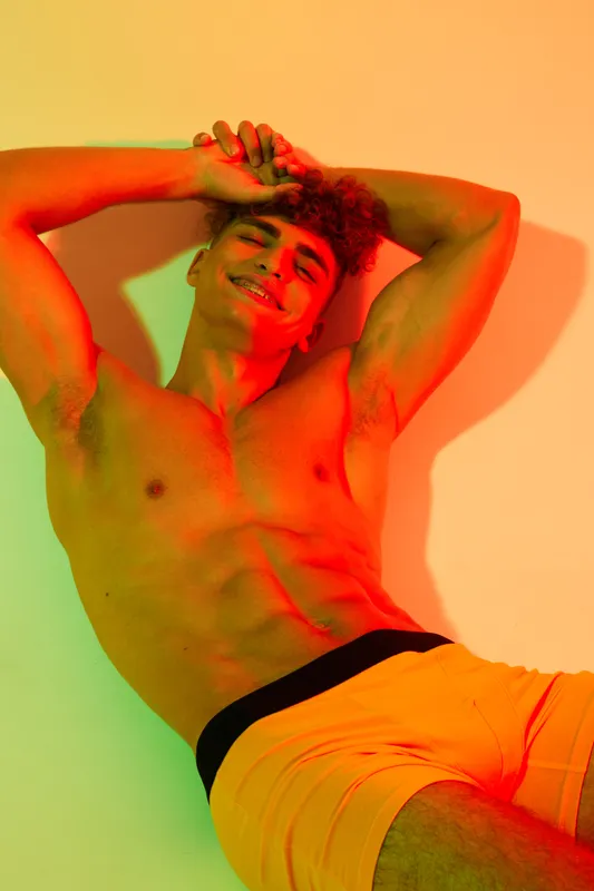 Young man with muscular body posing shirtless in colorful lighting, smiling and relaxed with arms behind head against plain background in studio portrait.