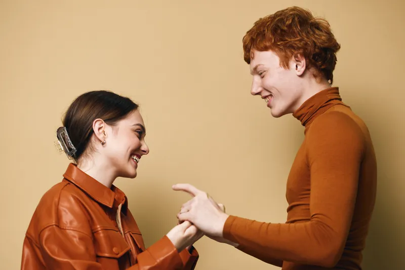 Young man and woman smiling and holding hands in casual clothing with warm expressions on beige background for friendship and connection.
