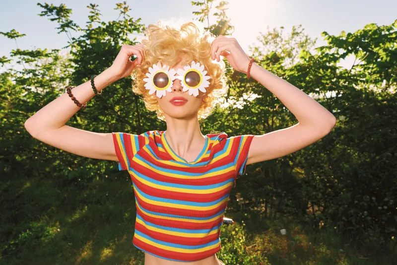 Playful woman outdoors with daisy sunglasses, striped rainbow top in bright sunlight, retro portrait mood emphasizing nature, garden and carefree lifestyle vibe, and bold color, soft focus, candid