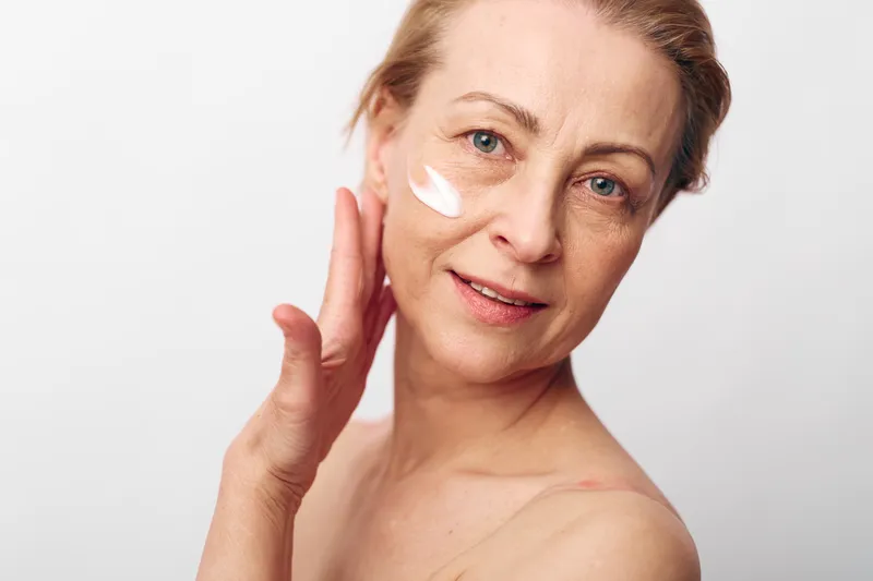 Mature woman applies white face cream on cheek with fingers, smiling gently. Close-up on light background. Concept of skincare, aging skin, self-care, and beauty routine.