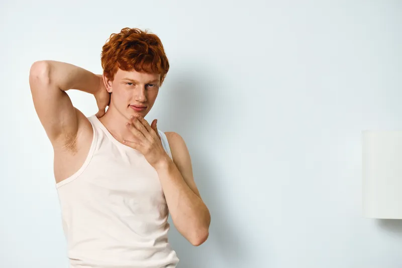 Young man with red hair in white tank top posing indoors with hand behind head and other hand near chin. Casual portrait in bright room with plain background.