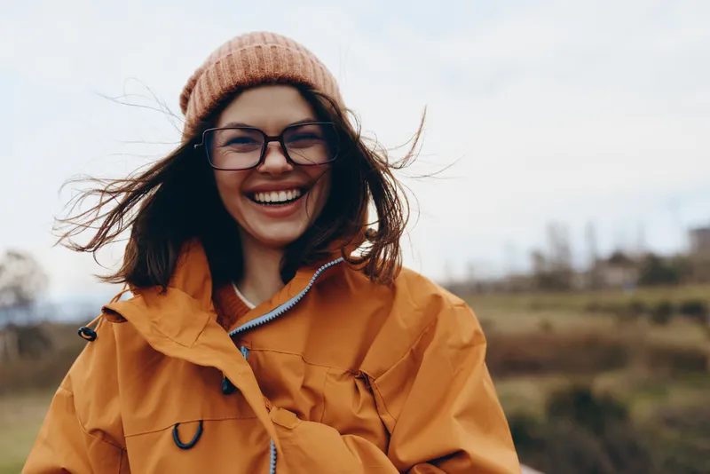 Woman smiling outdoors, lifestyle portrait in winter, wearing hat and glasses with orange jacket, wind lifting hair, warm coat and scarf, cheerful relaxed face, countryside field, background
