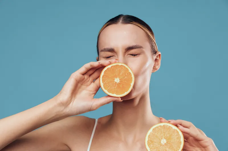 Young woman holds two halves of fresh orange fruit close to her face with closed eyes against blue background enjoying natural citrus aroma and healthy lifestyle concept.