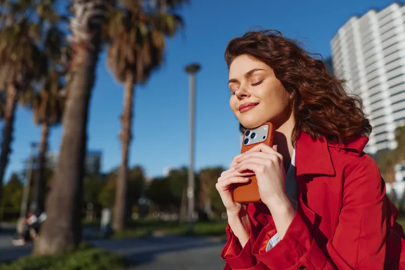 Young woman with curly hair enjoys sunlight outdoors while holding a smartphone close to her face with a peaceful smile.