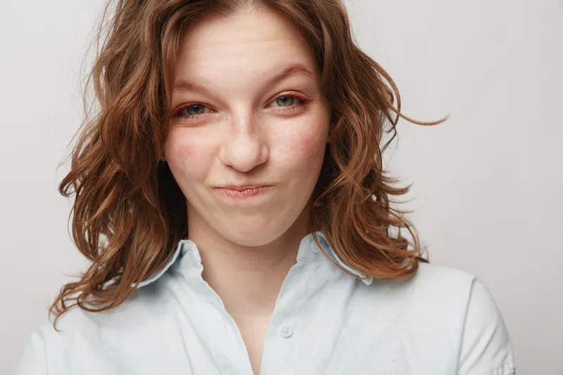 Young woman with curly hair makes funny face with pursed lips and squinted eyes. Casual white shirt and neutral background create simple, playful portrait concept of humor and personality.