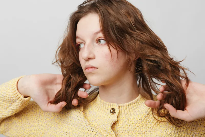 Young woman with long brown hair wearing yellow sweater looks sideways with thoughtful expression. Studio portrait on white background with natural lighting and casual style.