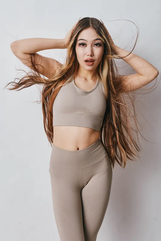 Young woman with long hair in fitness wear posing and touching head with hands indoors. Portrait, active lifestyle, energy and beauty in studio setting with neutral background.