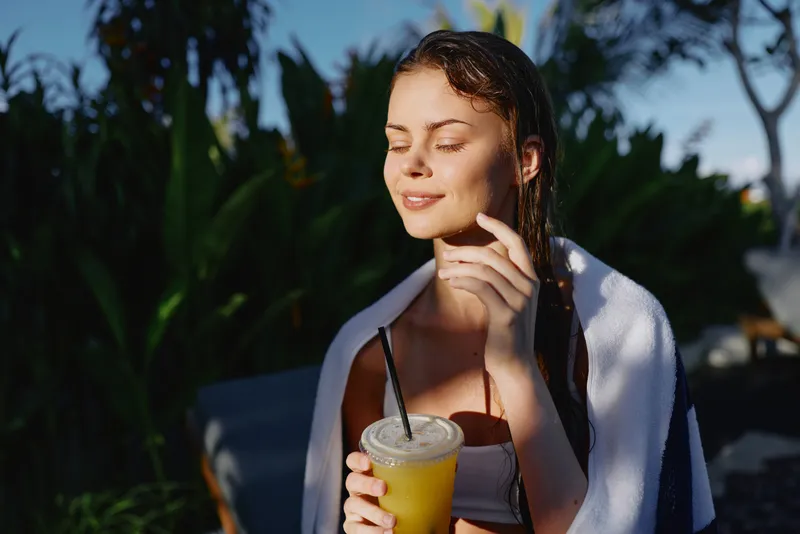 Young woman outdoors holding a cold drink with a straw, wrapped in a towel, enjoying sunlight with eyes closed, relaxed mood, tropical plants in background, casual lifestyle moment