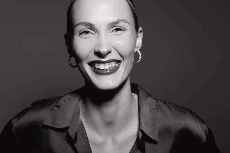 Smiling white woman with light skin and slicked back hair wears shiny shirt and hoop earrings in black and white studio portrait. Concept of happiness and beauty.