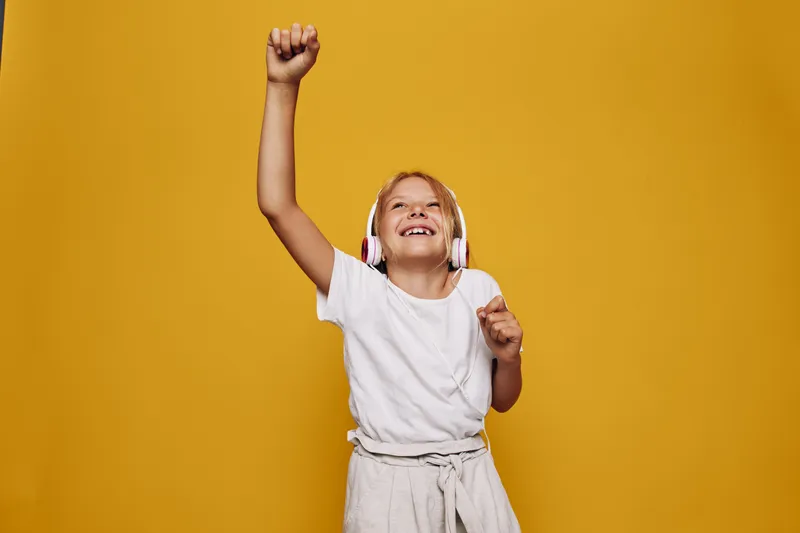 Happy child with headphones enjoys music and dances with raised arm on yellow background. Concept for joy, music, childhood, and fun activities.