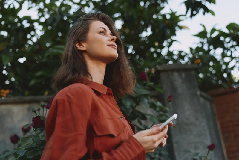 Woman in a red shirt stands outdoors, holding a smartphone, looking up with a calm smile, surrounded by greenery and blooming roses, lifestyle portrait
